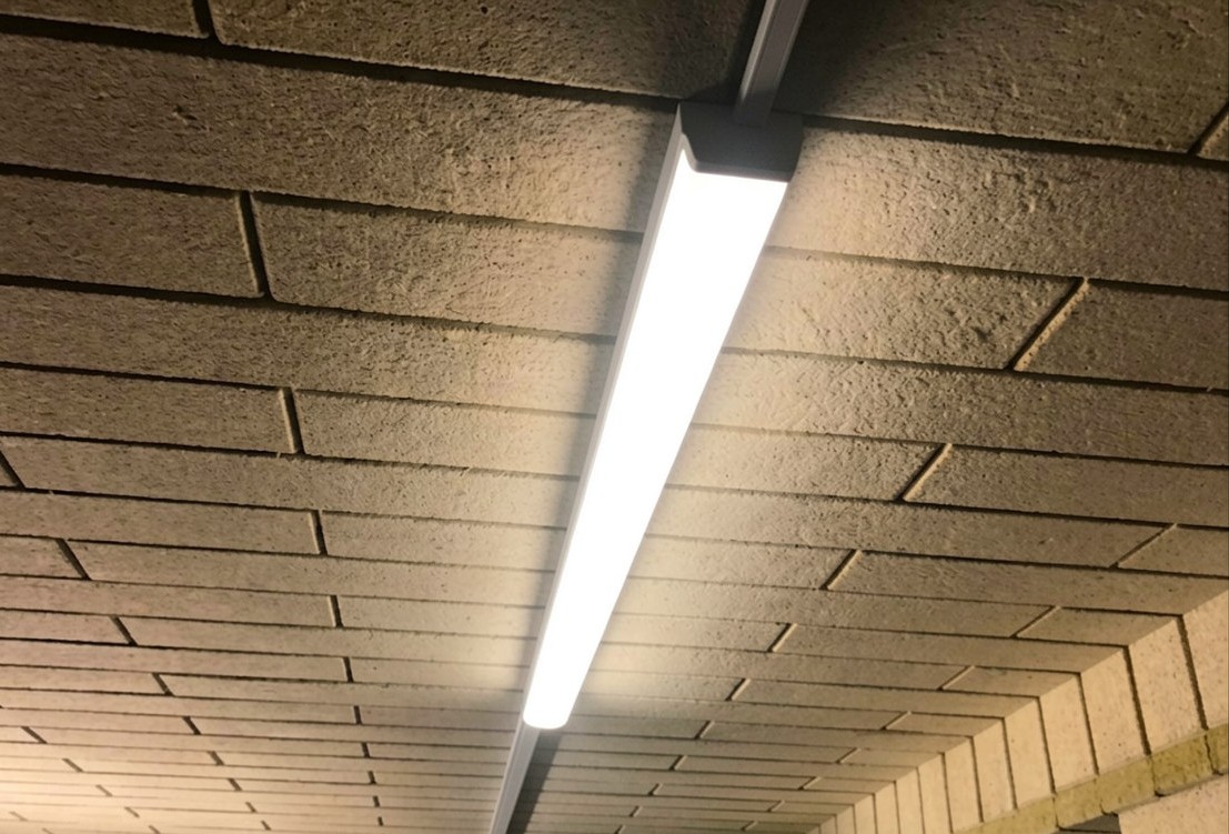 Detailní pohled na rozsvícené lineární LED svítidlo namontované na liště na strukturovaném stropě z izolačních lamelů. Perspektiva zdola nahoru zachycuje jas světla a texturu stropního materiálu.